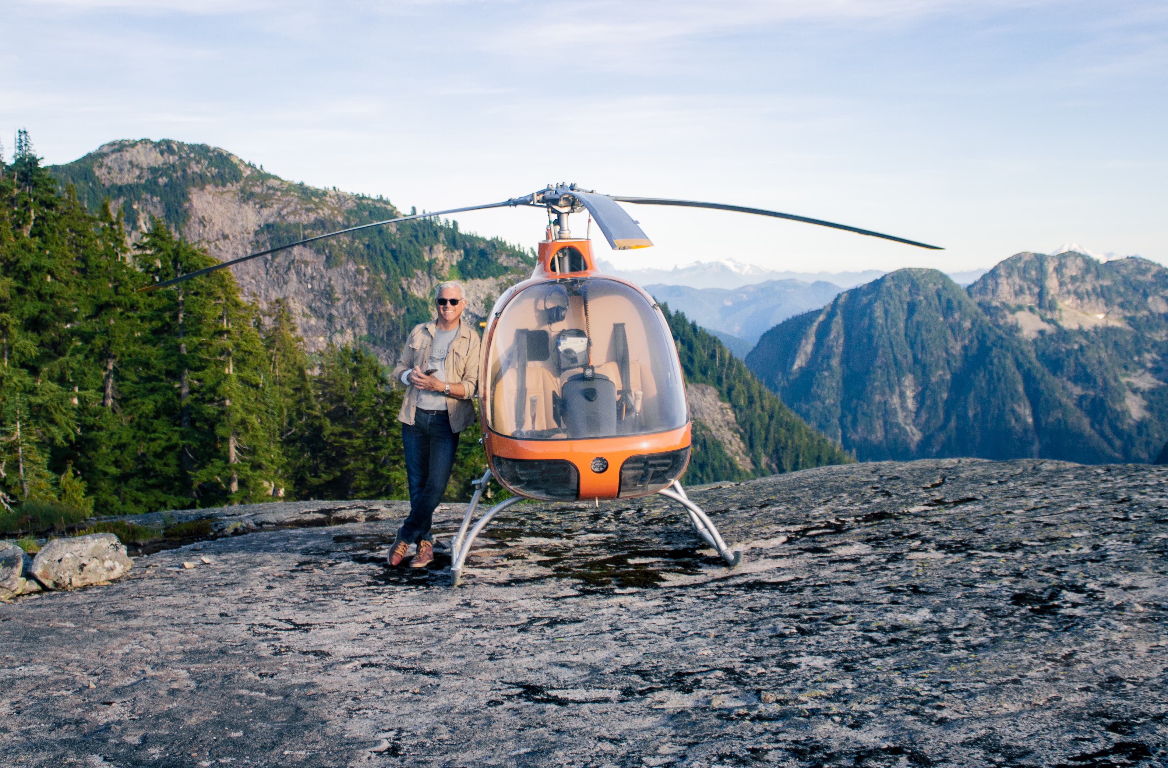 a close up photograph of a man standing next to a helicopter atop a mountain